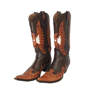 Könnte beinhalten: Ein Paar dunkelbraune Leder-Cowboystiefel mit spitzer Zehenpartie und wadenhohem Schaft. Die Stiefel sind mit orangefarbenen Lederapplikationen in Form von Adlerflügeln verziert, die durch ein kleines weißes Herz am Schaft akzentuiert werden.