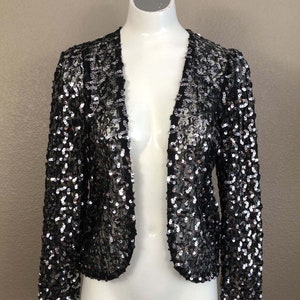 May include: A black sequined jacket with long sleeves and a deep V-neck. The jacket is made of a sheer fabric with silver sequins.