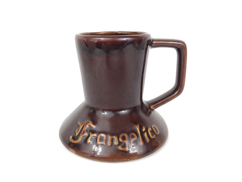 May include: A brown ceramic mug with a handle, featuring the word "Frangelico" in a lighter brown script. The mug has a unique shape, wider at the base and tapering towards the top.