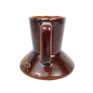May include: A brown ceramic mug with a handle. The mug has a flared base and a cylindrical body. The word "Nico" is written in gold lettering on the base. The mug is likely vintage and suitable for coffee or tea.