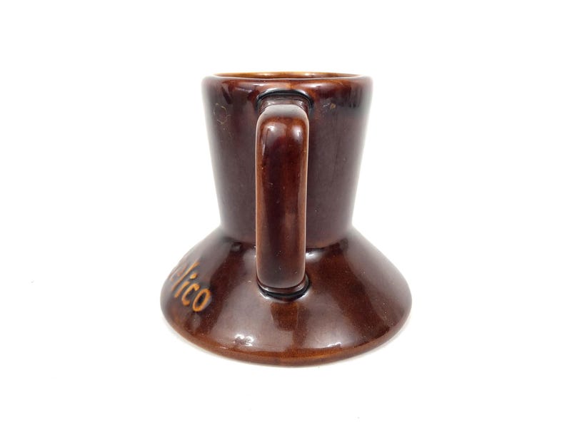 May include: A brown ceramic mug with a handle. The mug has a flared base and a cylindrical body. The word "Nico" is written in gold lettering on the base. The mug is likely vintage and suitable for coffee or tea.