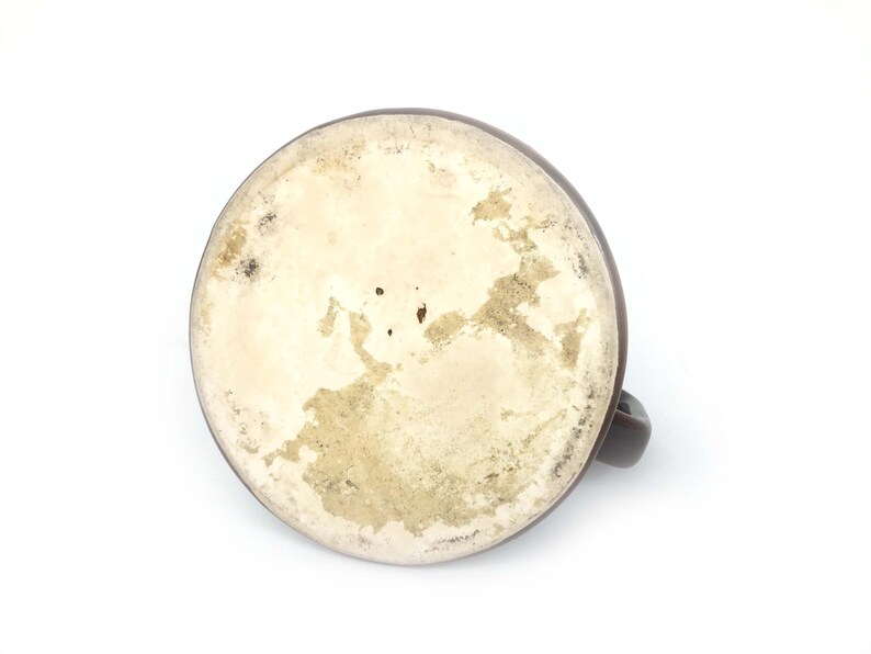 May include: A brown ceramic mug, seen from below, with a handle. The base interior is off-white with brown and tan staining. The mug is set against a white background.