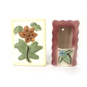 May include: Two ceramic wall planters, one with a yellow background and a green and orange flower design, the other with a pink background and a blue and green flower design.