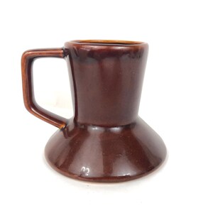 May include: A brown ceramic mug with an unusual hourglass shape. The mug has a handle and a glossy finish. The colour is a rich, dark brown, and the mug appears to be handmade. The mug is suitable for coffee, tea, or other beverages.