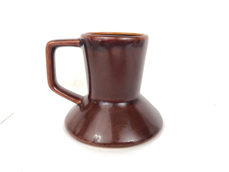 May include: A brown ceramic mug with an unusual hourglass shape. The mug has a handle and a glossy finish. The colour is a rich, dark brown, and the mug appears to be handmade. The mug is suitable for coffee, tea, or other beverages.