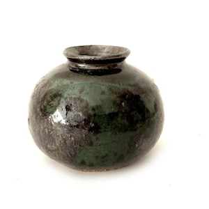 May include: A small, round ceramic vase with a dark green and black glaze. The vase has a wide body and a narrow neck. The surface has a textured appearance, and the vase is set against a white background.