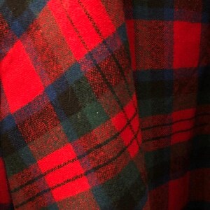 May include: A fabric with a plaid pattern featuring intersecting lines in vibrant red, deep forest green, and dark navy blue. The textile displays a woven texture with a repeating grid design that creates a classic tartan appearance across the surface.