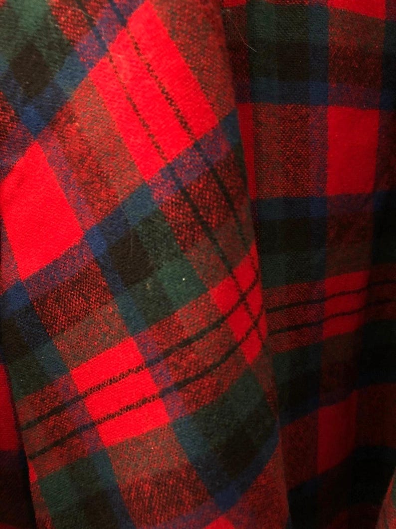 May include: A fabric with a plaid pattern featuring intersecting lines in vibrant red, deep forest green, and dark navy blue. The textile displays a woven texture with a repeating grid design that creates a classic tartan appearance across the surface.