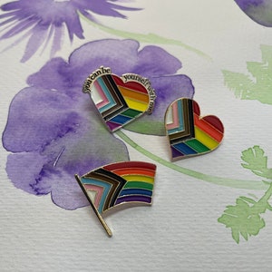May include: Three enamel pins featuring a rainbow pride flag design. The pins are in the shape of a heart and a flag. The heart pins have the text "you can be yourself with me" around the edge.