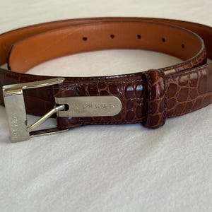 May include: A brown leather belt with a crocodile pattern. The belt has a silver buckle and a brown interior. The brand name "RALPH LAUREN" is visible on the buckle. The belt is a classic accessory.