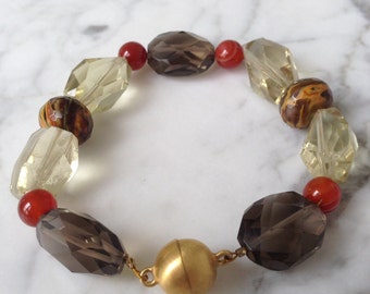 Può includere: Un braccialetto di perline con un mix di perline sfaccettate e opache nei toni del marrone, giallo e rosso. Il braccialetto ha una chiusura magnetica color oro. Le perline sono infilate con una catena color oro.