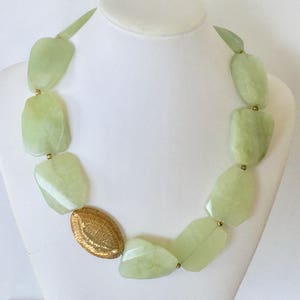 May include: A light green jade stone necklace with a gold oval pendant. The necklace is made of large, irregular shaped jade stones.