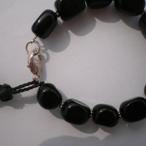 Pu&ograve; includere: Un bracciale con perline nere lisce e lucide separate da piccoli distanziatori in metallo color argento. Il bracciale include una chiusura a moschettone color argento e una nappa in filo nero attaccata all'anello metallico vicino.