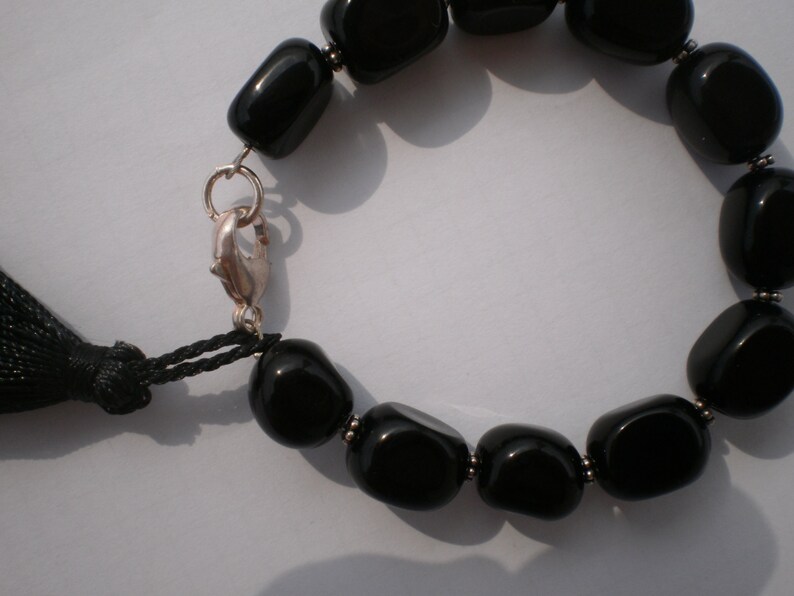 Pu&ograve; includere: Un bracciale con perline nere lisce e lucide separate da piccoli distanziatori in metallo color argento. Il bracciale include una chiusura a moschettone color argento e una nappa in filo nero attaccata all'anello metallico vicino.