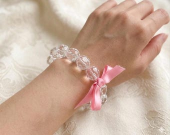 Può includere: Un braccialetto di perline trasparenti e sfaccettate con un fiocco di raso rosa. Il braccialetto è indossato su un polso, mettendo in risalto le qualità di cattura della luce delle perline. Il fiocco aggiunge un tocco delicato al gioiello.