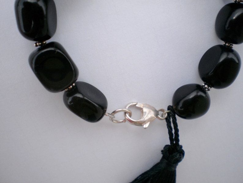 Pu&ograve; includere: Un bracciale con perline nere lisce e lucide, separate da piccoli distanziatori in metallo color argento. Il gioiello &egrave; fissato con una chiusura a moschettone color argento e presenta una nappa in filo nero vicino alla chiusura.