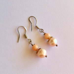 May include: A pair of dangle earrings featuring peach-colored pearls and smaller round beads. The earrings have silver-toned ear wires and decorative metal accents. The pearls have a lustrous sheen, and the design is simple and elegant.