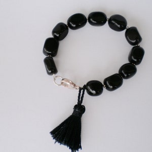 Pu&ograve; includere: Un bracciale composto da perline ovali nere lisce e lucide, separate da piccoli distanziatori color argento. Il bracciale presenta una chiusura a moschettone color argento e una nappa in filo nero appesa al meccanismo di chiusura.
