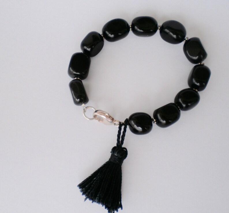 Pu&ograve; includere: Un bracciale composto da perline ovali nere lisce e lucide, separate da piccoli distanziatori color argento. Il bracciale presenta una chiusura a moschettone color argento e una nappa in filo nero appesa al meccanismo di chiusura.