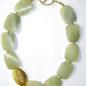 May include: A necklace featuring large, faceted, pale green stone beads strung together with small gold-toned spacer beads. The piece includes one oval, textured gold-toned metal bead and secures with a gold-toned hook and eye clasp.