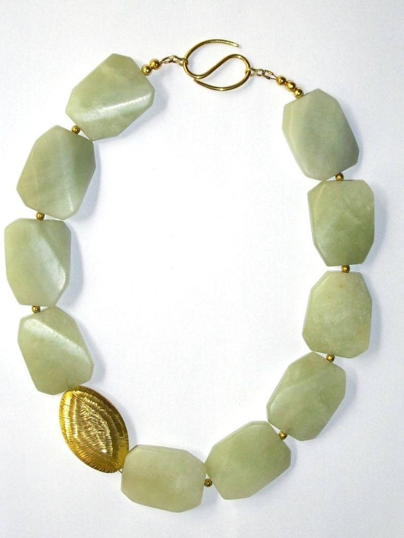 May include: A necklace featuring large, faceted, pale green stone beads strung together with small gold-toned spacer beads. The piece includes one oval, textured gold-toned metal bead and secures with a gold-toned hook and eye clasp.