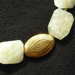 May include: A necklace featuring alternating pale green, faceted, irregular stone beads and a single, central, oval-shaped gold-toned metal bead with a textured, hammered surface finish, connected by small, round, gold-toned spacer beads.