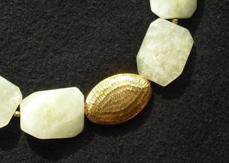 May include: A necklace featuring alternating pale green, faceted, irregular stone beads and a single, central, oval-shaped gold-toned metal bead with a textured, hammered surface finish, connected by small, round, gold-toned spacer beads.