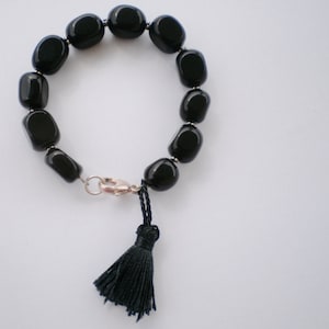 Pu&ograve; includere: Un bracciale composto da perline ovali nere, lisce e lucide, separate da piccoli distanziatori in metallo argentato. Il bracciale presenta una chiusura a moschettone argentata e una nappa in filo nero appesa al meccanismo di chiusura.