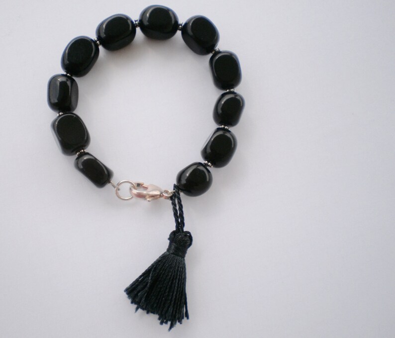 Pu&ograve; includere: Un bracciale composto da perline ovali nere, lisce e lucide, separate da piccoli distanziatori in metallo argentato. Il bracciale presenta una chiusura a moschettone argentata e una nappa in filo nero appesa al meccanismo di chiusura.