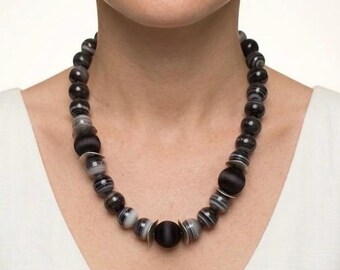May include: A beaded necklace featuring alternating round black matte beads and spherical beads with grey and white horizontal banding, accented by small silver-toned metal discs between the beads. The necklace is worn around the neck.
