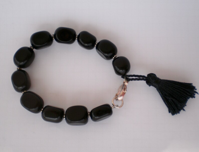 Pu&ograve; includere: Un braccialetto composto da perline rettangolari nere lisce e lucide, separate da piccoli distanziatori in metallo color argento. Il braccialetto presenta una chiusura a moschettone color argento e una nappa in filo nero attaccata alla chiusura.