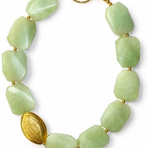 May include: A necklace featuring large, faceted, pale green stone beads strung together with small gold-toned metal spacers. One oval, textured gold-toned metal bead is positioned on the left side, and the piece closes with a gold-toned hook clasp.