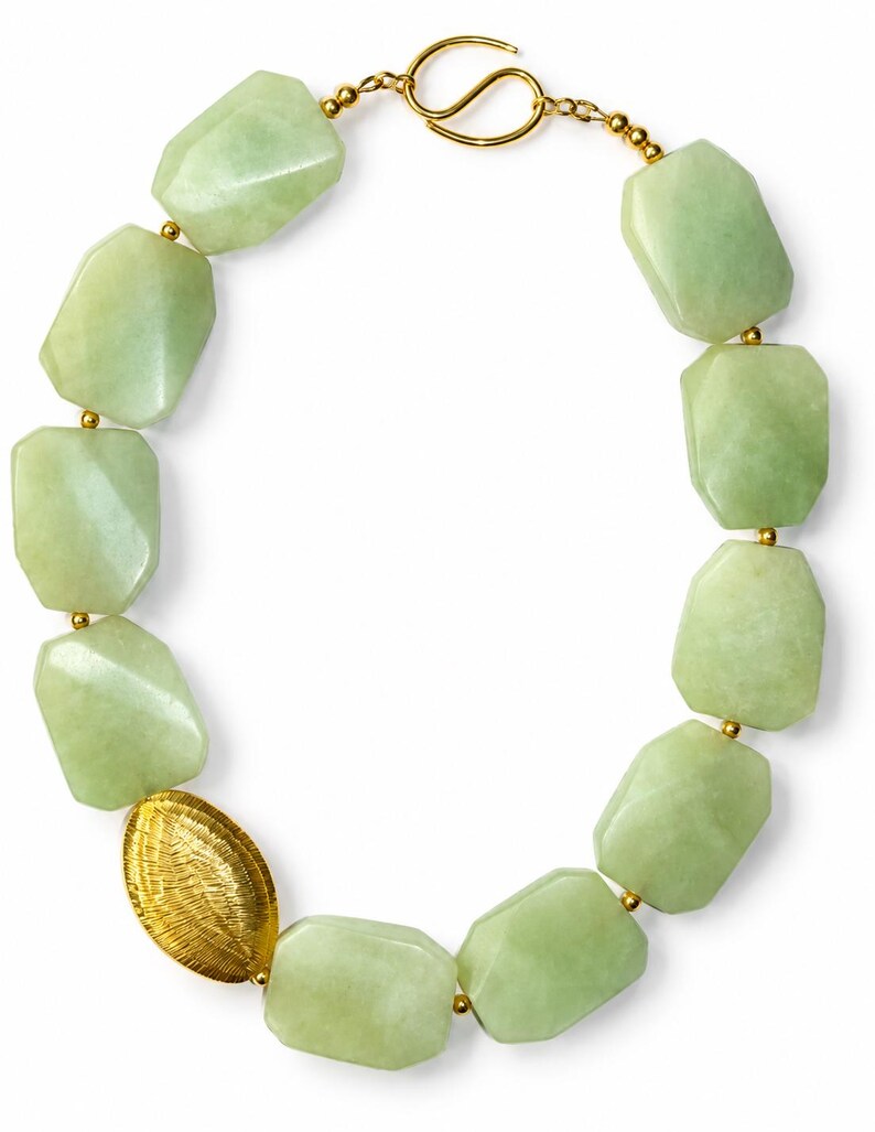 May include: A necklace featuring large, faceted, pale green stone beads strung together with small gold-toned metal spacers. One oval, textured gold-toned metal bead is positioned on the left side, and the piece closes with a gold-toned hook clasp.