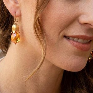 May include: A gold-toned drop earring featuring a faceted, amber-colored crystal bead suspended from a metal cap shaped like a flower bud. The earring hangs from a simple gold-toned hook, showcasing a clear, reflective surface and warm tones.