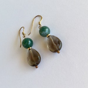 May include: A pair of dangle earrings featuring gold-colored hooks. Each earring has a green, round bead above a teardrop-shaped, smoky quartz bead. Small gold-colored beads accent the design. The earrings are displayed on a white surface.