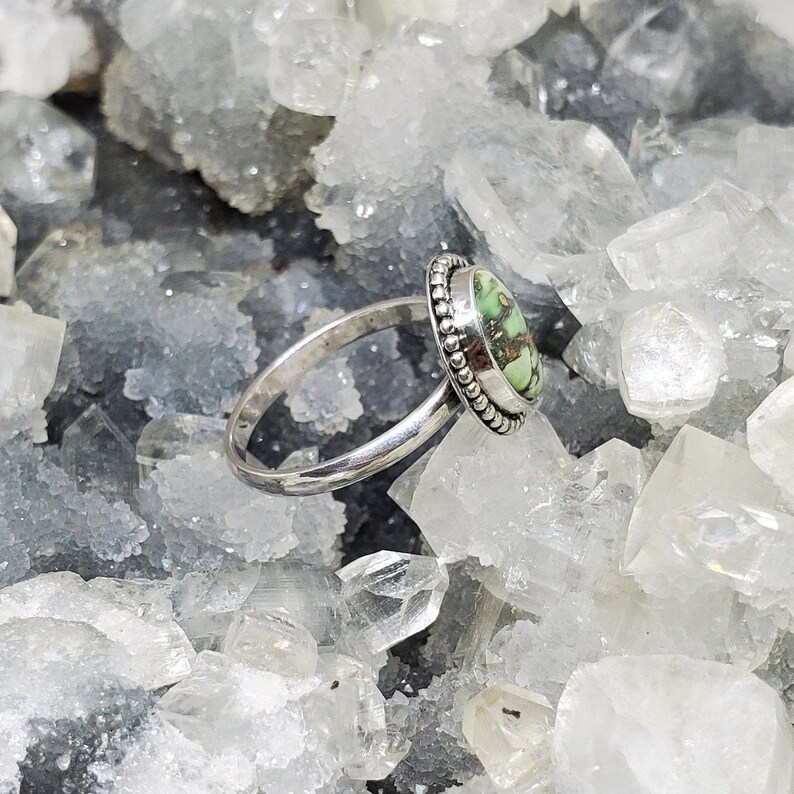 K&ouml;nnte beinhalten: Ein silberner Ring mit einem ovalen, gr&uuml;n-braunen Edelstein, der in einer Perlenfassung sitzt. Der Ring ist auf einem Bett aus wei&szlig;en Kristallen fotografiert.