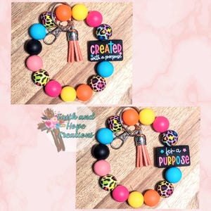 May include: A colorful wristlet keychain with a variety of round beads in shades of pink, yellow, orange, blue, black, and leopard print. The keychain includes a silver clasp, a leather tassel, and a black rectangle with the words "created with a purpose".