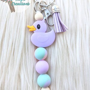 May include: Purple rubber duck keychain with a wooden bead and three pastel colored beads. The keychain has a silver key ring and a purple tassel.
