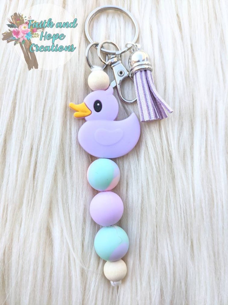 May include: Purple rubber duck keychain with a wooden bead and three pastel colored beads. The keychain has a silver key ring and a purple tassel.