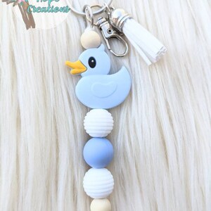 May include: A blue rubber duck keychain with a white tassel and wooden beads. The beads are white, blue, and beige.