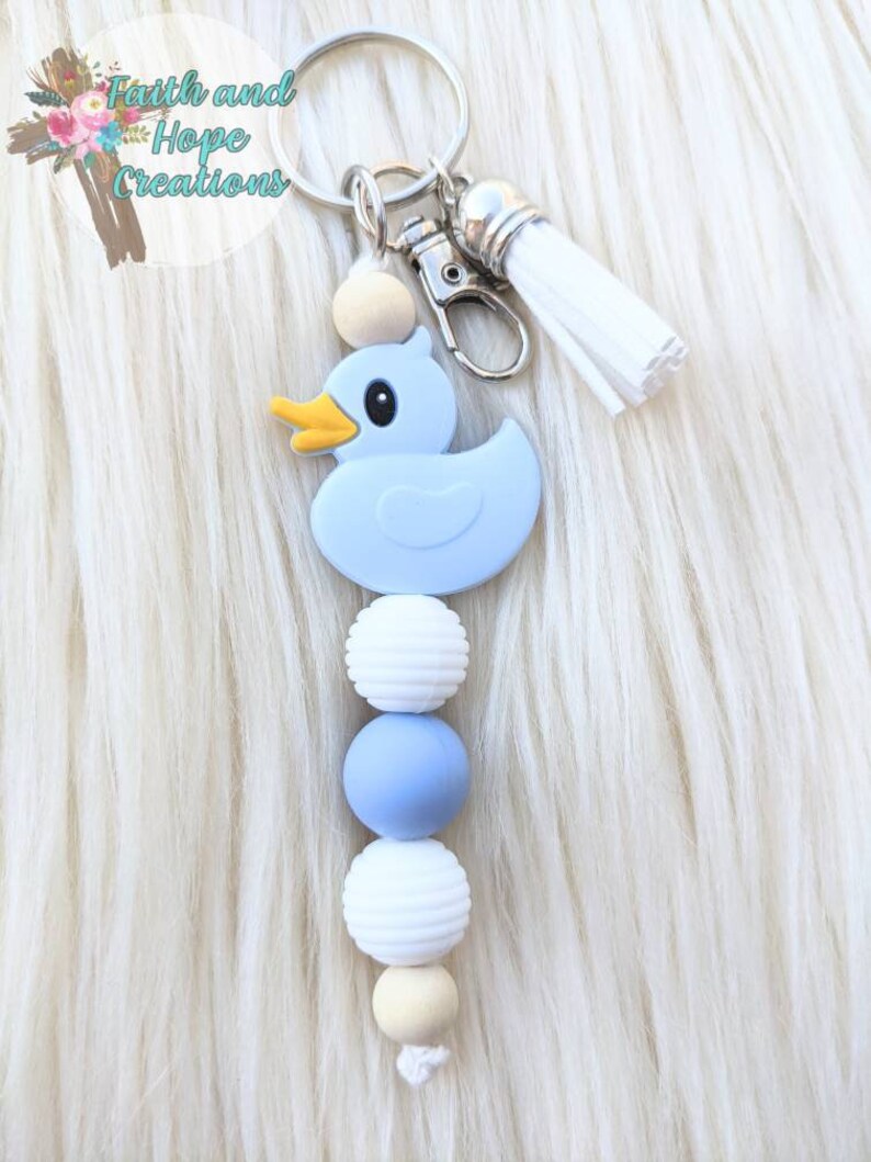 May include: A blue rubber duck keychain with a white tassel and wooden beads. The beads are white, blue, and beige.