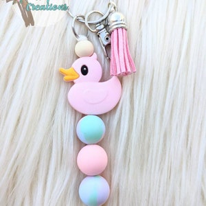 May include: Pink rubber ducky keychain with a pink tassel and wooden beads. The beads are alternating colors of pink, blue, and white.