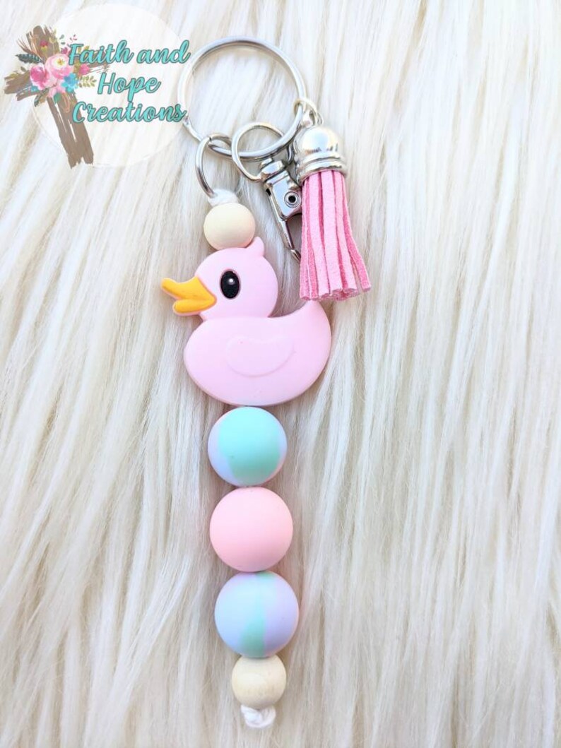 May include: Pink rubber ducky keychain with a pink tassel and wooden beads. The beads are alternating colors of pink, blue, and white.