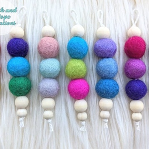 May include: Eight rainbow-colored felt ball garlands with wooden beads. Each garland has four felt balls and one wooden bead. The felt balls are red, orange, yellow, green, blue, purple, pink, and gray.