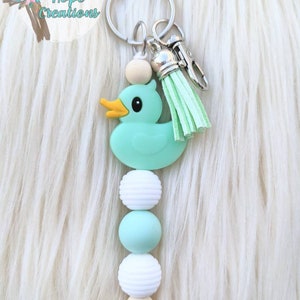 May include: A teal rubber duck keychain with a mint green tassel and a string of white and teal beads. The keychain is attached to a silver key ring.