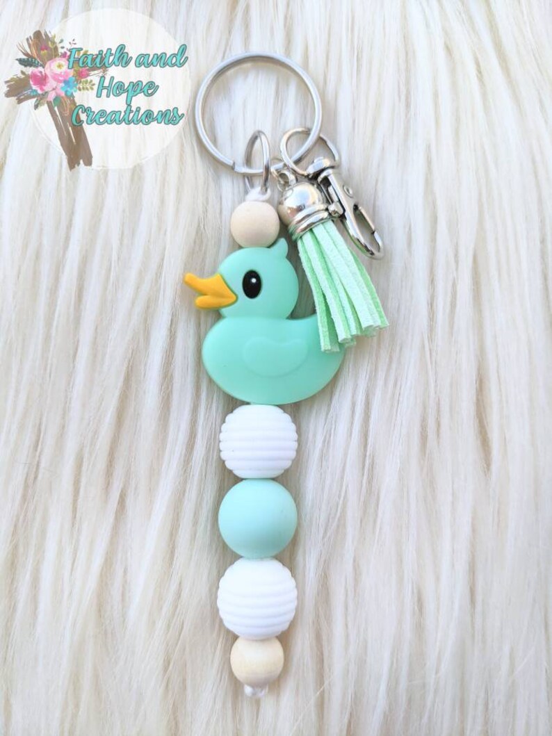 May include: A teal rubber duck keychain with a mint green tassel and a string of white and teal beads. The keychain is attached to a silver key ring.
