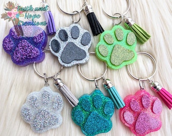 May include: A set of six colorful glitter paw print keychains with silver key rings and tassels. The paw prints are made of resin and have a variety of colors, including purple, gray, green, white, blue, and pink.