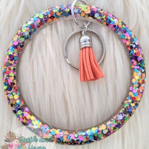 May include: A clear, glitter-filled bangle keychain with a silver keyring and a coral tassel. The bangle is filled with colorful confetti-like glitter. The keychain is a fashionable accessory.