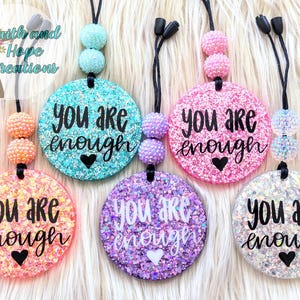 May include: Glittery, round car charms in various colors, including pink, teal, and purple. Each charm has the phrase "YOU ARE ENOUGH" in black script, with a heart symbol. The charms have beaded accents and black cords.