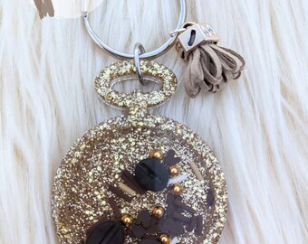 May include: Gold glitter keychain with a round, clear resin charm filled with coffee beans, sprinkles, and chocolate pieces. The keychain has a silver ring and a brown tassel.